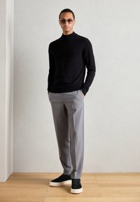 Maglione a collo alto nero in maglia, pantaloni grigi su misura e sneakers nere con suola bianca. Il modello si trova su un pavimento in legno chiaro contro uno sfondo grigio.