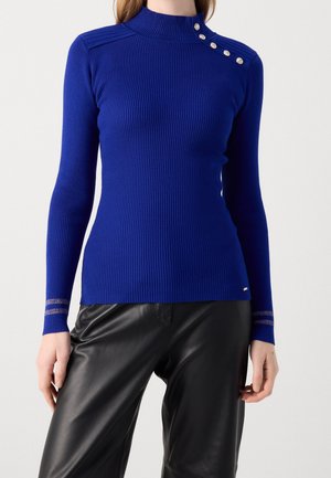 Pull côtelé bleu cobalt ajusté avec des boutons argentés sur l'épaule et des rayures argentées aux poignets, associé à un pantalon en cuir noir.