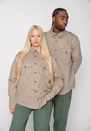 Una mujer rubia y un hombre de piel oscura están de pie uno al lado del otro, vestidos con camisas beige abotonadas y pantalones verdes, sobre un fondo claro y liso.