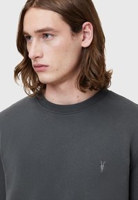 Sudadera gris oscuro con cuello redondo de canalé, que presenta un pequeño logotipo bordado de skull de cabra en el lado izquierdo del pecho. Textura suave.