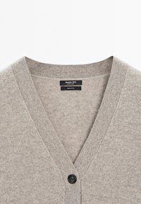 Ljusgrå bomullskardigan med V-hals, med en enda svart knappknäppning och en mjuk, texturerad yta. Varumärkesetikett synlig inuti.