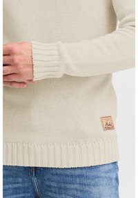 Pulover tricotat de culoare bej deschis cu manșete și tiv cu aspect côté, având o mică patch din material maro cu logo în partea stângă jos.