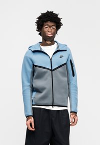 Φούτερ με φερμουάρ Nike σε μπλε και γκρι, με κομψό σχέδιο, μαύρες λεπτομέρειες, πλαϊνή τσέπη με φερμουάρ και υφασμάτινη υφή.