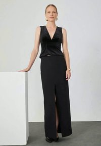 Black velvet sleeveless top with V-neckline paired with a long black skirt featuring a thigh-high slit. Model stands beside a white pedestal.