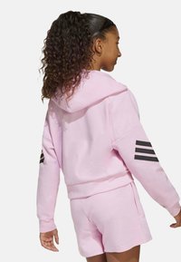 Sudadera rosa de corte relajado con un diseño de tres franjas negras en las mangas, combinada con unos pantalones cortos rosa a juego. Tejido suave y texturizado.