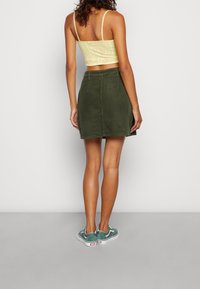 Olivgrön kordflossekjol med bakfickor, kombinerad med en ljusgul, mönstrad crop top och gröna sneakers med vita accenter.