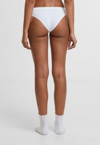 Baltās bikini apakšbikses ar gludu apdari, vidēju segumu un zemu jostasvietu aizmugurē, pārī ar baltām viļņotām potīšu zeķēm.