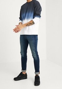 Felpa ombré che sfuma dal blu scuro al bianco, abbinata a jeans di denim scuro e sneakers nere. Presenta maniche lunghe e polsini arrotolati.