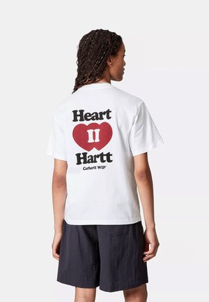 Osoba z długimi warkoczami, ubrana w białą koszulkę z czerwonym sercem i czarnym napisem "Heart II Hartt" oraz czarnymi teksturowanymi szortami, zwrócona plecami.