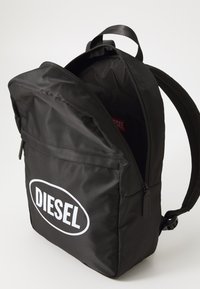 Juodas nailono kuprinė su užtrauktuku, turinti ryškų baltą "DIESEL" logotipą. Įtraukti paminkštinti petnešos ir šoninis kišeni.