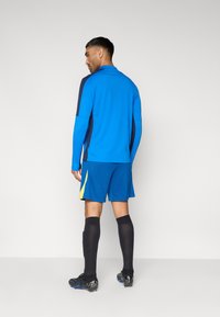 Μπλε μπλουζάκι με μακριά μανίκια και σκούρες μπλε λεπτομέρειες, συνδυασμένο με μπλε σορτς με κίτρινη πλαϊνή ρίγα. Μαύρες αθλητικές κάλτσες και παπούτσια με τάπες.