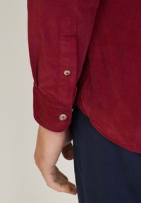 Camicia di velluto a coste rossa con polsini a bottoni, una texture sottile e un orlo arrotondato, abbinata a pantaloni blu scuro.