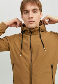 Veste zippée marron avec un col haut, des cordons noirs réglables, et des poches latérales zippées. Tissu lisse avec une finition mate.