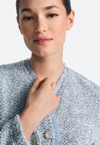 Világoskék tweed zakó fodros gallérral, kerek ezüst gombbal és texturált anyaggal, amely finom szőtt mintákkal van kiemelve.