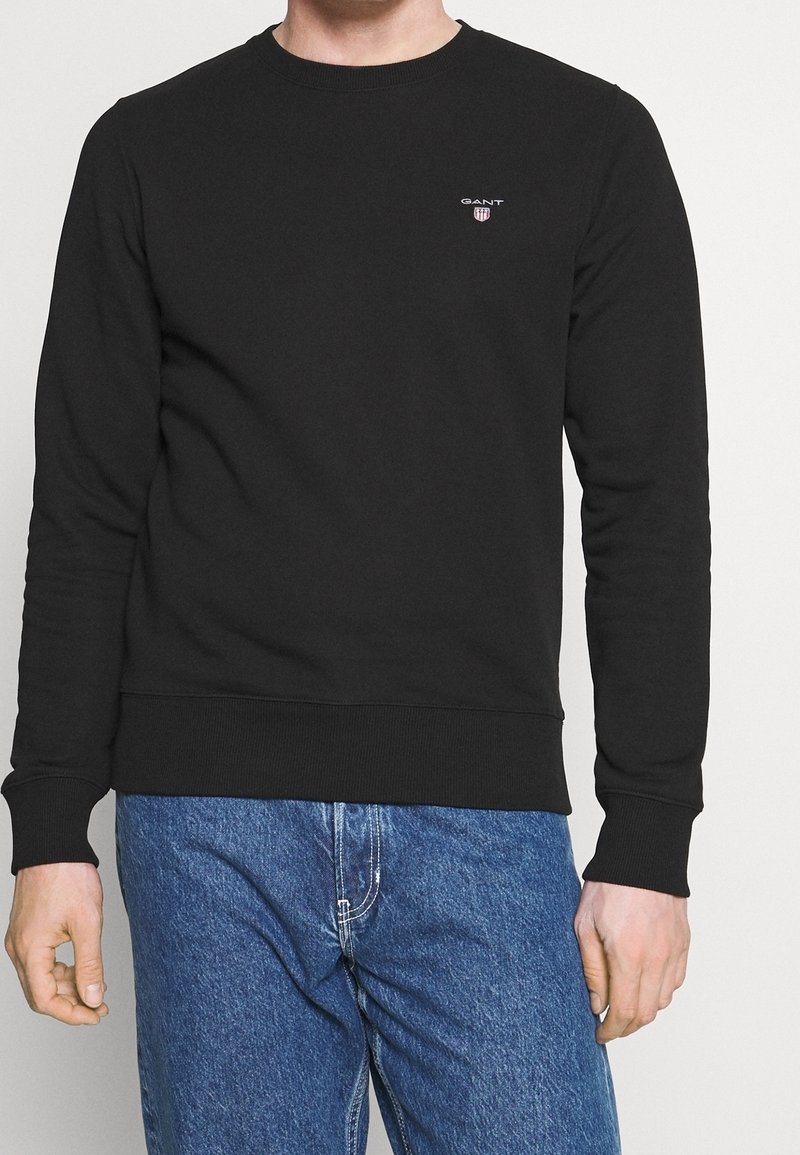 Felpa nera in cotone con scollo tondo, polsini e vita a costine. Presenta un piccolo logo sul petto. Indossata con jeans in denim blu.