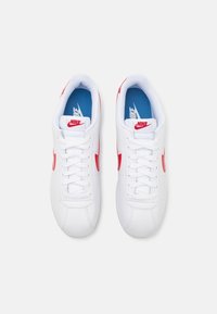 White Nike sneakers with red accents and textured synthetic upper. Features a lace-up design, a light blue insole, and logo detailing.