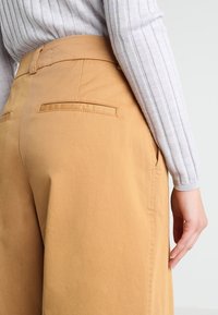 Ljusbruna, midjehöga byxor i mjukt tyg med bakficka och en strukturerad silhuett, kombinerade med en grå ribbad topp.