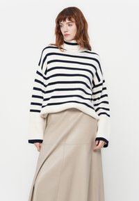Maglione a righe in crema e blu marina con collo alto. Vestibilità ampia, polsini a coste e texture liscia. Accompagnato da una gonna beige neutra.