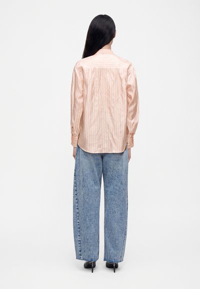 Femme aux longs cheveux noirs, de dos, portant une chemise rose clair à rayures, un jean large bleu et des chaussures à talons hauts noires.