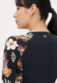 Lange mouw zwemtop in zwart met een bloemenpatroon in bruin- en crèmekleuren. Voorzien van een ronde hals en een logo-accent op de achterkant.