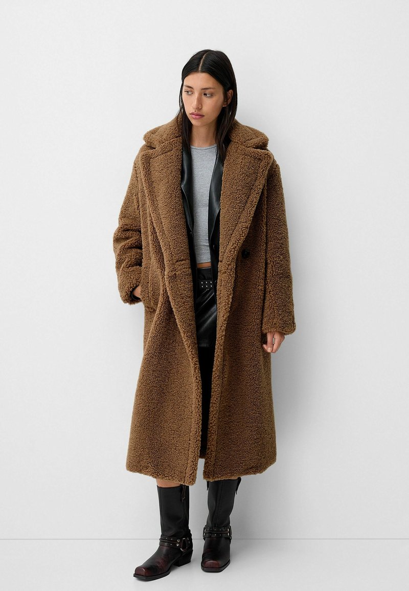 Manteau teddy oversized marron avec une texture douce, design double boutonnage, grands revers et poches latérales, superposé sur un haut gris et un pantalon noir.