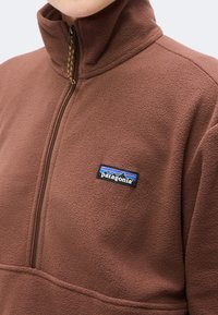 Кафяв флисов пуловер с четвърт цип и лого на Patagonia от лявата страна на гърдите, носен от човек, изрязан на нивото на врата.