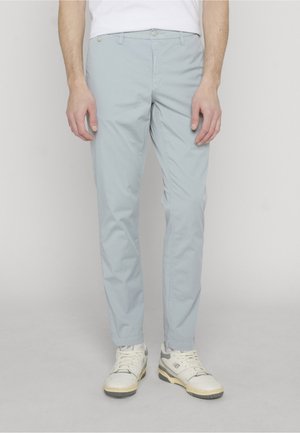 Homme portant un pantalon slim bleu clair, des baskets blanches et une chemise blanche sur un fond uni clair.