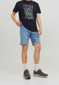 Man wearing navy graphic t-shirt, light blue rolled denim shorts, white socks, and gray sneakers standing against plain background.