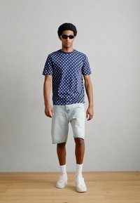 Μπλε ναυτικό μπλουζάκι με πουά, ανοιχτό τζιν σορτς, λευκά αθλητικά παπούτσια και γυαλιά ηλίου. Το μοντέλο έχει αφρο μαλλί και στέκεται σε ξύλινο δάπεδο.