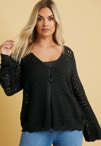 Schwarze Strickjacke mit strukturiertem offenen Gewebe, V-Ausschnitt, Knöpfen vorne und Glockenärmeln. Getragen über einem enganliegenden Oberteil und kombiniert mit Jeans.