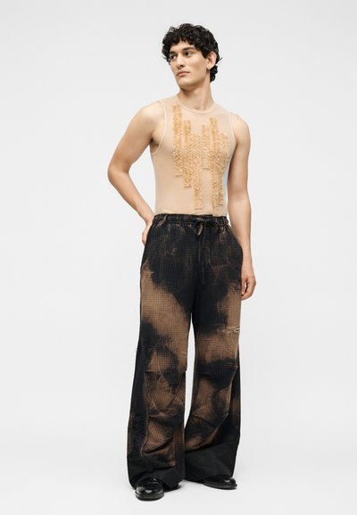 Άνδρας που φοράει αμάνικη μπεζ μπλούζα με υφή σε κάθετες ρίγες, χαλαρά μαύρα και καφέ παντελόνια με τύπωμα tie-dye και μαύρα παπούτσια, στέκεται με το ένα χέρι πίσω.