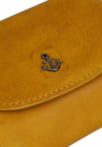 Billetera amarilla de cuero con forma redondeada, con un emblema de ancla de metal y costuras en contraste. Textura suave con un ligero brillo.