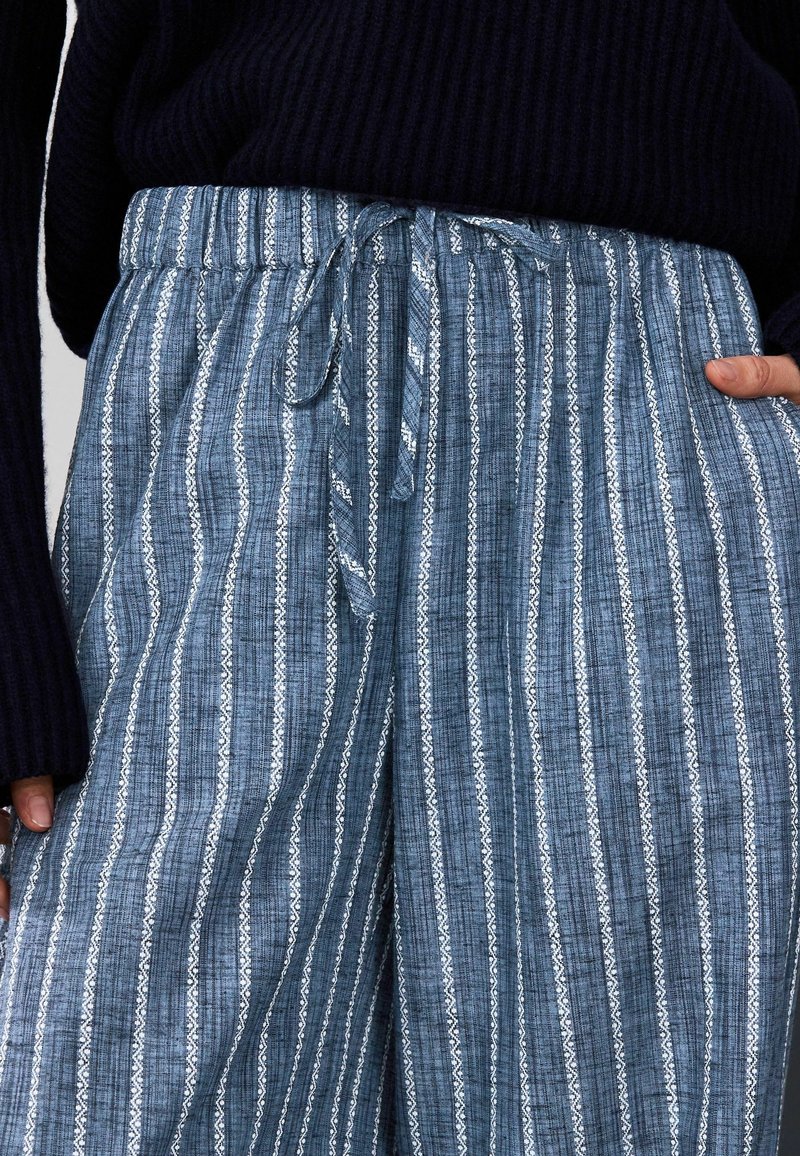 Niebieskie szerokie spodnie w wzory z troczkiem w pasie i pionowymi paskami z misternymi białymi wzorami. Teksturowana tkanina dodaje głębi.