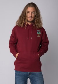 Bordeauxkleurige hoodie van sweatstof. Voorzien van een buidelzak en een capuchon met trekkoord. Op de linkerkant van de borst staat een kleurrijk grafisch ontwerp.