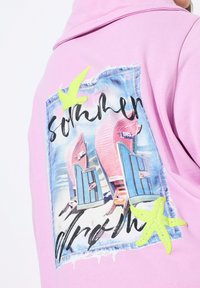 Sudadera rosa con un gráfico en la parte posterior que muestra sillas de playa, un fondo azul y acentos de estrellas de mar en verde neón.