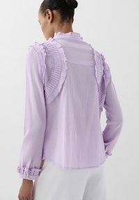 Paarse, lichte blouse met gerimpelde accenten langs de schouders en manchetten, textiel met structuur en een iets losse pasvorm.