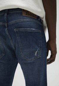 Person som bär mörkblå denimjeans med ett litet hål vid bakfickan och en synlig brun varumärkesetikett på midjebandet.