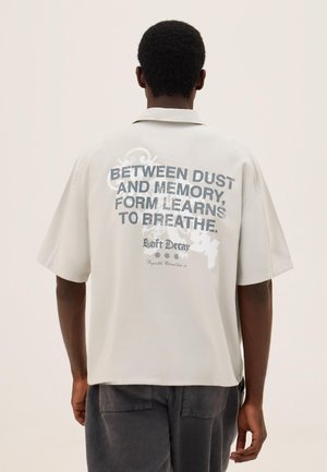 Mand med kort krøllet hår iført en lysebeige kortærmet skjorte med teksten "Between dust and memory, form learns to breathe" på ryggen.