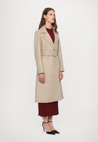 Bežo spalvos tranšėjos paltas su plačiu apykakle ir dirželiu liemens srityje, pasižymintis lygiu paviršiumi. Dėvimas ant bordo suknelės ir derinamas su juodais aukštakulniais.