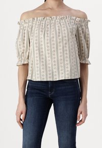 Top décolleté en crème avec un motif floral bleu et des rayures verticales. Présente un col volanté et des manches bouffantes. Porté avec un jean sombre.