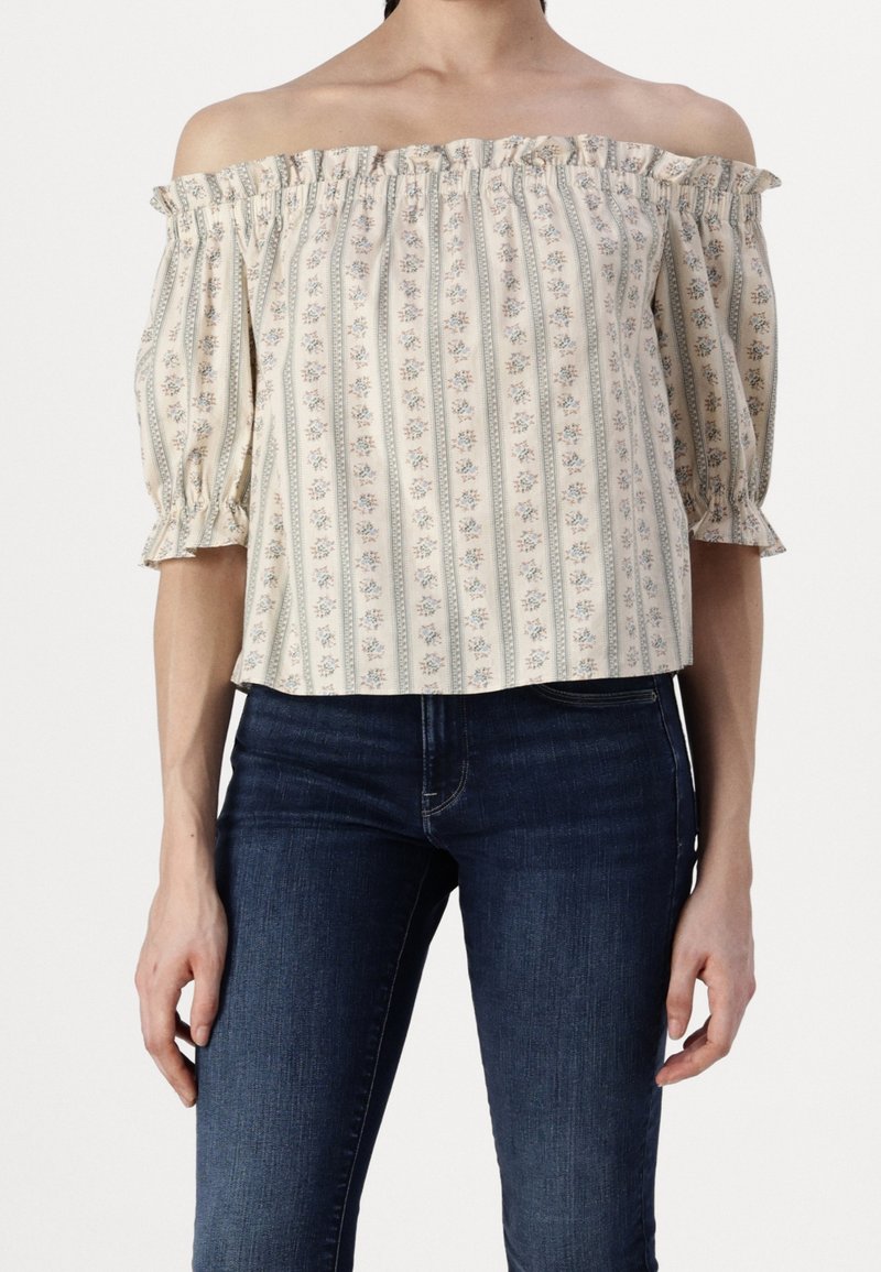 Top décolleté en crème avec un motif floral bleu et des rayures verticales. Présente un col volanté et des manches bouffantes. Porté avec un jean sombre.