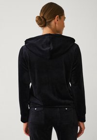 Zwarte, zachte velours hoodie met een kangoeroezak en capuchon. Lange mouwen en een ontspannen pasvorm. Soepele textuur met minimale hardware accenten.