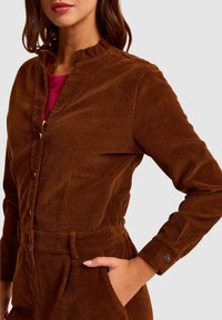 Brązowa koszula z korduroyu z falbaną na kołnierzu, zapięcie na guziki i długie rękawy. Posiada kieszeń oraz teksturowany materiał w pionowe paski.