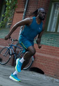Homme courant dans une chemise sans manches teal et un short teal avec des accents lime. Il porte des chaussures de course bleu vif avec des semelles blanches. Fond en brique.