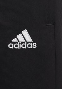 Biały haftowany logo Adidas z trzema paskami i tekstem na czarnym teksturowanym tle.