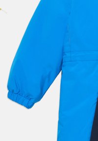 Μπλε αδιάβροχο μπουφάν με μανίκι και ελαστικό μανίκι, που διαθέτει λεία υφή και αντίθετο σκούρο πάνελ κατά μήκος της πλευράς.