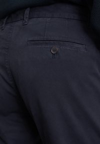 Dunkelmarineblaue Hose aus einem glatten Stoff. Verfügt über eine Gesäßtasche mit einem einzigen braunen Knopf und dezenten Nähdetails.