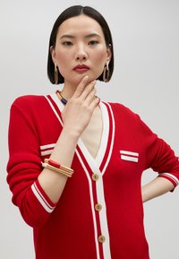 Red ribbed cardigan with white accents, V-neckline, and four gold buttons. Features a striped design detail on the left side.