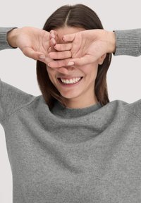 Grauer Pullover mit runder Ausschnitt und gerippten Bündchen. Der Stoff wirkt weich und leicht strukturiert. Die Hände des Models bedecken sein Gesicht.