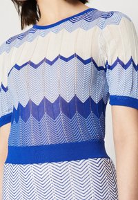 Robe en maille présentant un motif géométrique en zigzag bleu et blanc, avec une finition texturée et une ceinture unie bleue. Manches courtes.